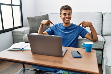 Young handsome hispanic man using laptop sitting on the floor looking confident with smile on face, pointing oneself with fingers proud and happy.