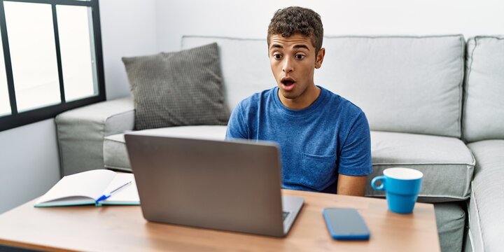 Young handsome hispanic man using laptop sitting on the floor afraid and shocked with surprise expression, fear and excited face.