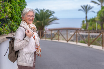 Smiling caucasian elderly woman walking in outdoors between palm trees and sea. Senior carefree female holding backpack and looking at camera