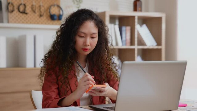 Designer wearing a red suit works with computer notebook to check the work before sending to customer. A freelance woman writes down important messages in a post-it note to remind her not to forget.