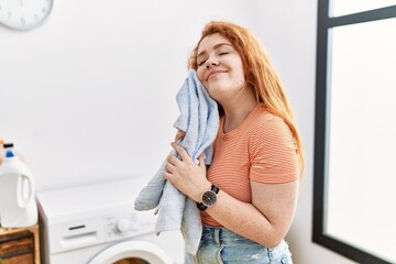 Young redhead woman smiling confident touching face with soft clothes at laundry room