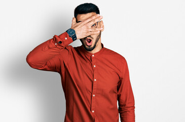 Young hispanic man with beard wearing business shirt and glasses peeking in shock covering face and eyes with hand, looking through fingers with embarrassed expression.