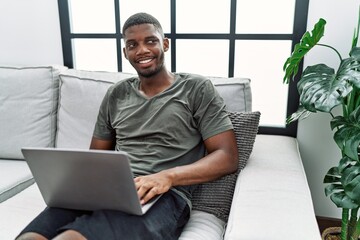 Young african american man using laptop at home sitting on the sofa looking away to side with smile on face, natural expression. laughing confident.