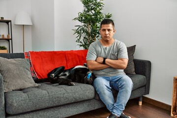 Young latin man and dog sitting on the sofa at home skeptic and nervous, disapproving expression on face with crossed arms. negative person.