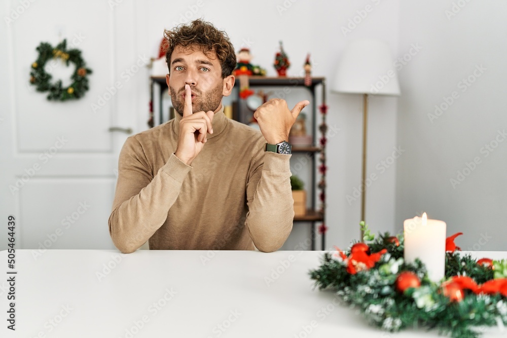 Sticker young handsome man with beard sitting on the table by christmas decoration asking to be quiet with f