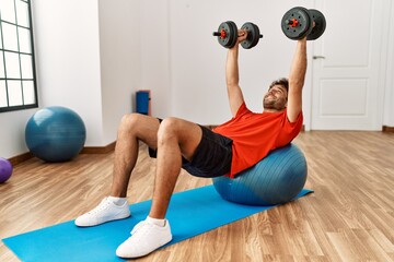 Young hispanic man smiling confident training using dumbbells and fit ball at sport center
