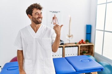 Young handsome physiotherapist man working at pain recovery clinic smiling with happy face looking and pointing to the side with thumb up.