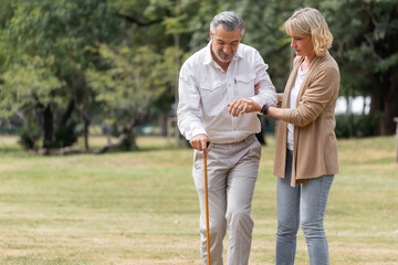 Caucasian senior woman supporting sick senior man with walking cane at the public park. Elderly couple helping each other. Retirement health and illness concept