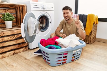 Young handsome man putting dirty laundry into washing machine smiling with happy face winking at...
