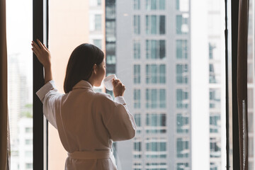 Thai woman wearing a bath robe drinking coffee and admiring the scenery in a hotel room in morning