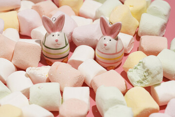 Two bright figurines of easter bunnies surrounded by white fluffy marshmallows on a pink surface, a close up