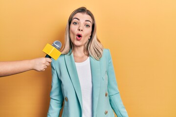 Beautiful caucasian woman being interviewed by reporter holding microphone scared and amazed with...