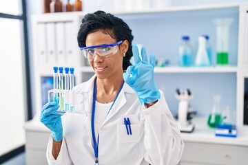 Middle age hispanic woman working at scientist laboratory doing ok sign with fingers, smiling friendly gesturing excellent symbol