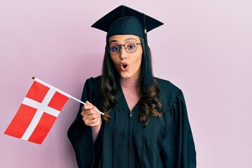 Young hispanic woman wearing graduation uniform holding denmark flag scared and amazed with open mouth for surprise, disbelief face
