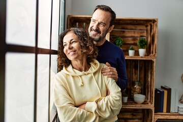 Middle age hispanic couple smiling happy leaning on the window at home.