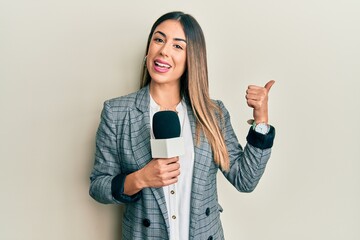 Young hispanic woman holding reporter microphone pointing thumb up to the side smiling happy with...