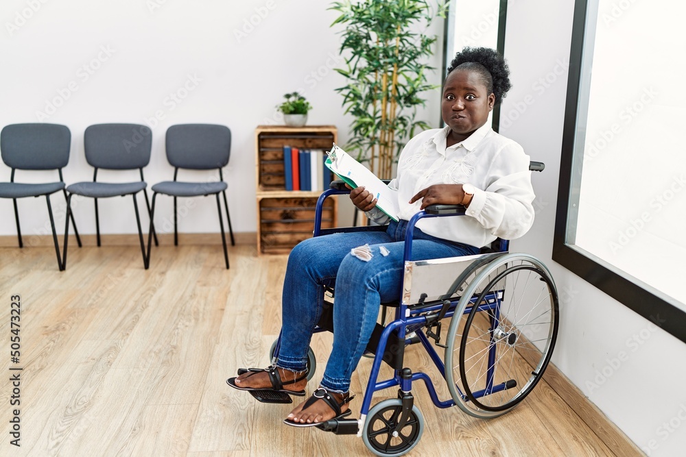 Poster young black woman sitting on wheelchair at waiting room puffing cheeks with funny face. mouth inflat