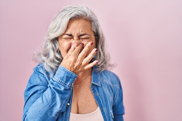 Middle age woman with grey hair standing over pink background smelling something stinky and disgusting, intolerable smell, holding breath with fingers on nose. bad smell - obrazy, fototapety, plakaty
