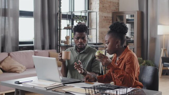 Waist-up of young African American couple sitting at desk in living room at daytime, making online purchase via computer, using credit card, woman eating apple