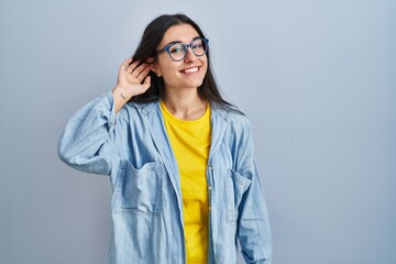 Young hispanic woman standing over blue background smiling with hand over ear listening an hearing to rumor or gossip. deafness concept.