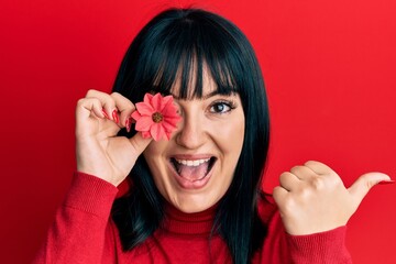 Young hispanic woman holding sunflower over eye pointing thumb up to the side smiling happy with open mouth