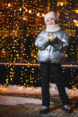 Girl with tangerines in her hands with Christmas lights. Smiling girl in warm clothes holding mandarin in hands and looking at the camera at night with blurry lights. Holidays theme