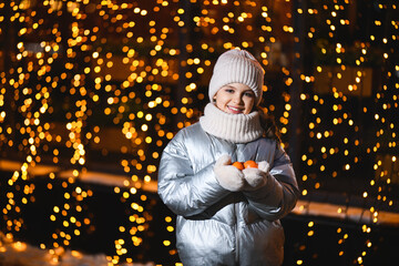 Girl with tangerines in her hands with Christmas lights. Smiling girl in warm clothes holding mandarin in hands and looking at the camera at night with blurry lights. Holidays theme