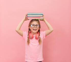 Little girl in eyeglasses and headphones with stack of books on her head on pink background. Back...