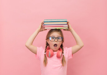 Little scared girl in eyeglasses and headphones with stack of books on her head with mouth open in...