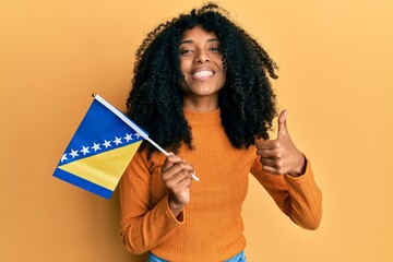 African american woman with afro hair holding bosnia herzegovina flag smiling happy and positive,...