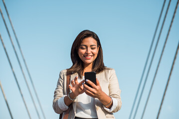 A happy elegant young latin woman stands and uses her phone to type an important message. A businesswoman using technologies outdoors. - Powered by Adobe