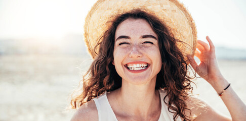 Young joyful woman in white shirt wearing hat smiling at camera on the beach - Traveler girl...