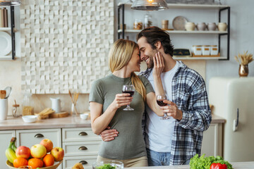 Glad caucasian millennial guy with stubble kisses blond wife with glass of wine