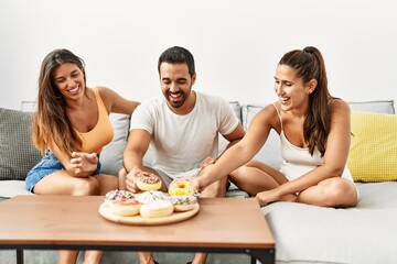 Three hispanic friends smiling happy eating doughnut at home.