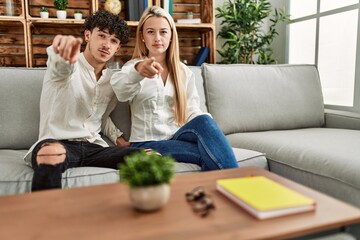 Young beautiful couple sitting on the sofa at home pointing with finger to the camera and to you, confident gesture looking serious