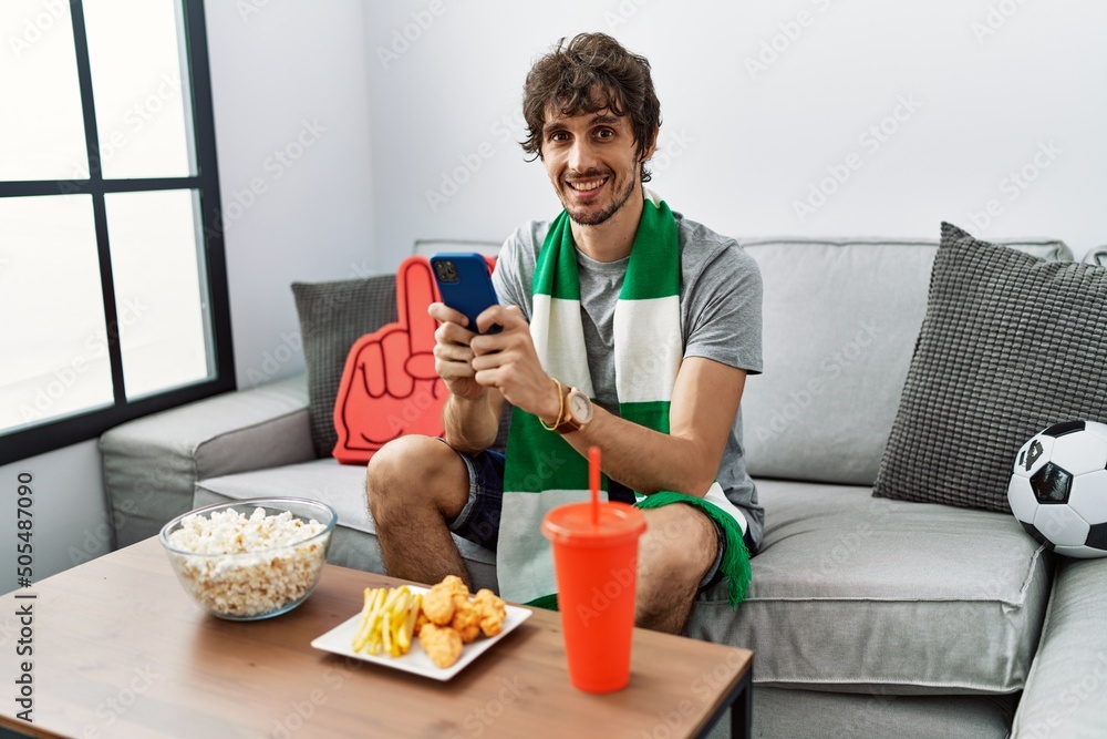 Sticker Young hispanic man using smartphone supporting soccer match at home