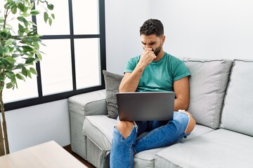 Young handsome man with beard using computer laptop sitting on the sofa at home smelling something...