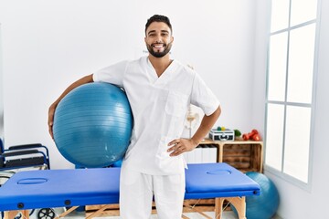 Young arab man wearing physiotherapist uniform holding fit ball at clinic