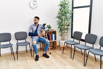 Young arab man sitting on chair with arm sling holding crutch at clinic waiting room