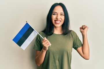 Young hispanic girl holding estonia flag screaming proud, celebrating victory and success very...