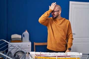 Young bald man with beard hanging clothes at clothesline surprised with hand on head for mistake,...