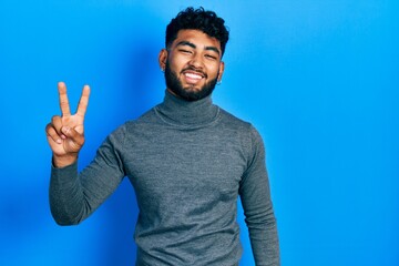 Arab man with beard wearing turtleneck sweater smiling looking to the camera showing fingers doing victory sign. number two.