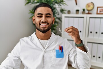 Young indian man working at dentist clinic holding invisible aligner looking positive and happy standing and smiling with a confident smile showing teeth