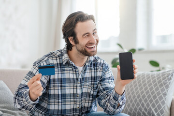 Satisfied young caucasian male with stubble winks sit on sofa with credit card and phone with empty screen