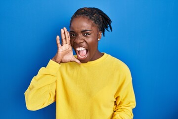 Beautiful black woman standing over blue background shouting and screaming loud to side with hand on mouth. communication concept.