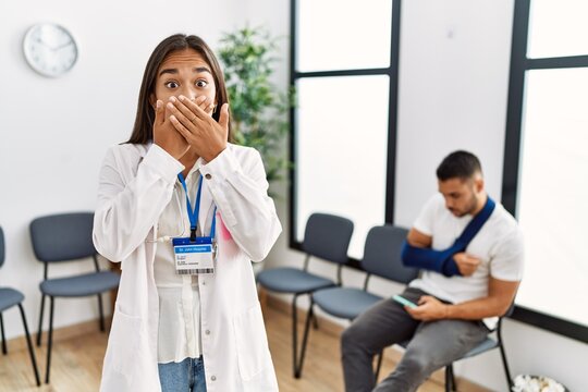 Young asian doctor woman at waiting room with a man with a broken arm shocked covering mouth with hands for mistake. secret concept.