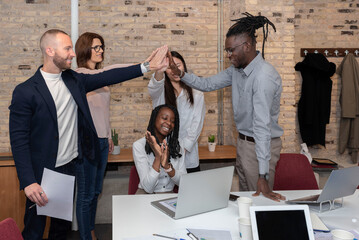  multiracial entrepreneurs in the conference room achieve corporate success by celebrating market leadership by clapping their hands around an African-American female colleague seated at the table. 