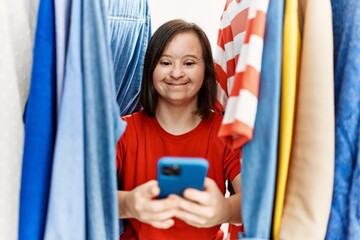 Brunette woman with down syndrome standing between hangers using smartphone at retail shop