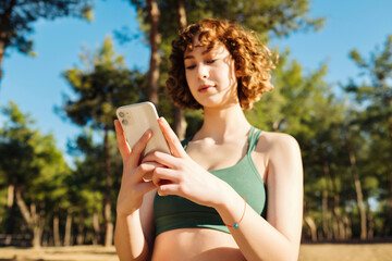 Athletic young redhead woman wearing green sports bra using her smart phone while standing on a city park, outdoors. Watching video or messaging with friends or scrolling on social media.