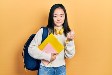 Young chinese girl holding student backpack and books doing money gesture with hands, asking for...